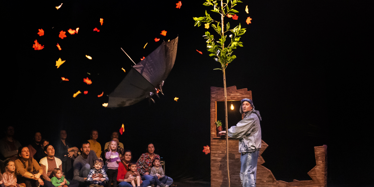 je ziet een deel van het publiek en het podium met de acteur die naast een boompje staat en een paraplu die weg vliegt met herfstbladeren in de lucht
